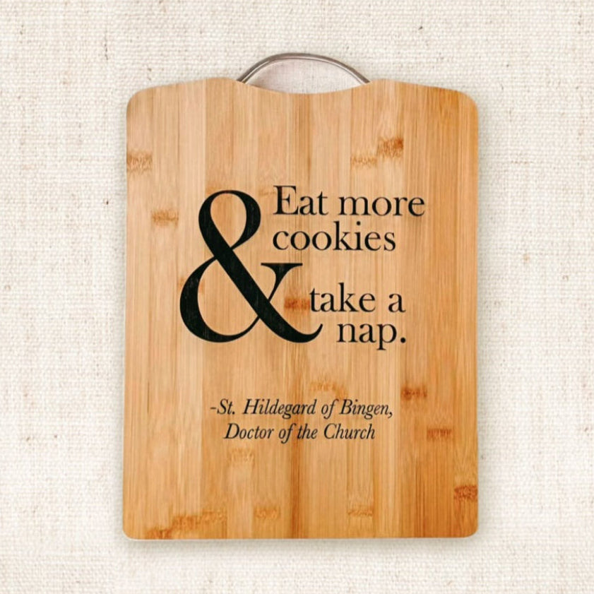 bamboo cutting board with fictitious quote of Saint Hildegard "Eat more cookies and take a nap" on a light canvas background