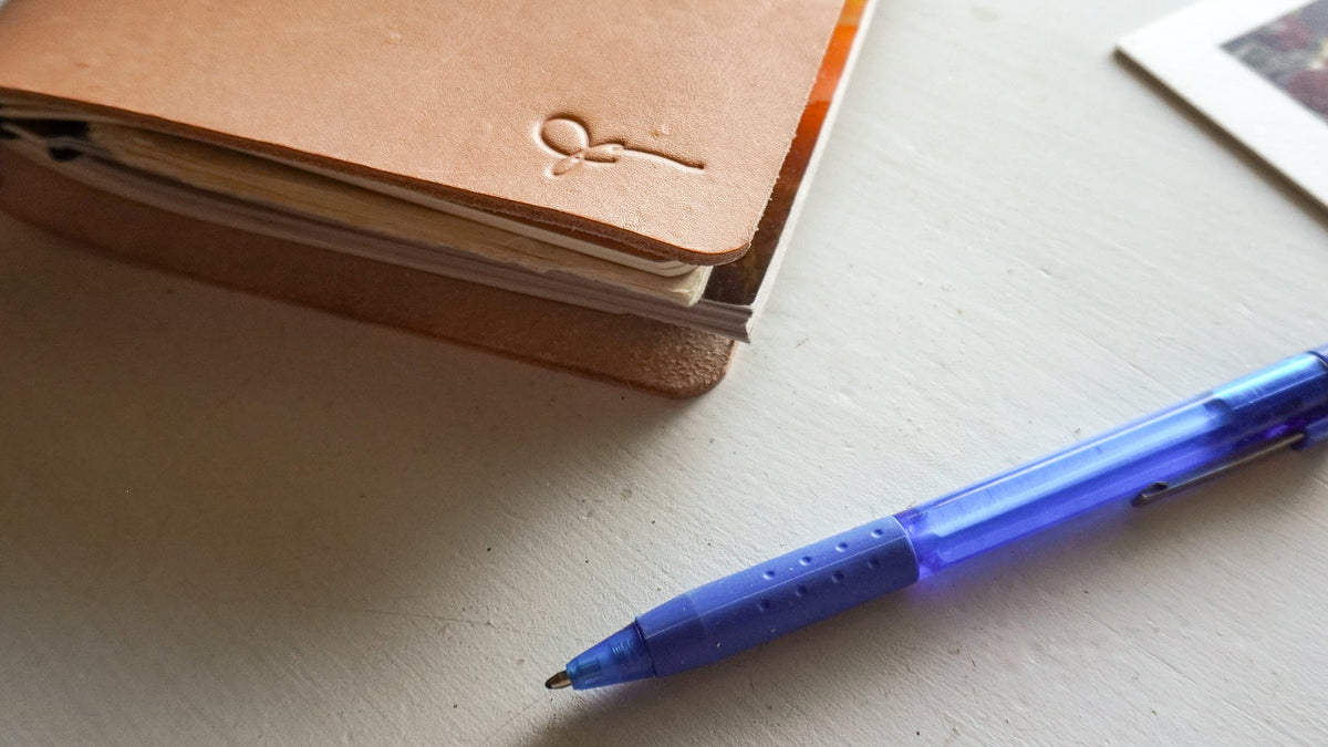 A natural leather notebook with black elastic closure and embossed signature on the front cover on a white table next to a blue pen