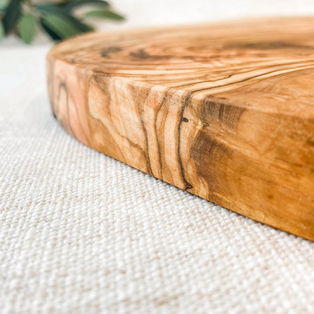 A close up of an olive wood cutting board on white canvas with an olive branch in the background